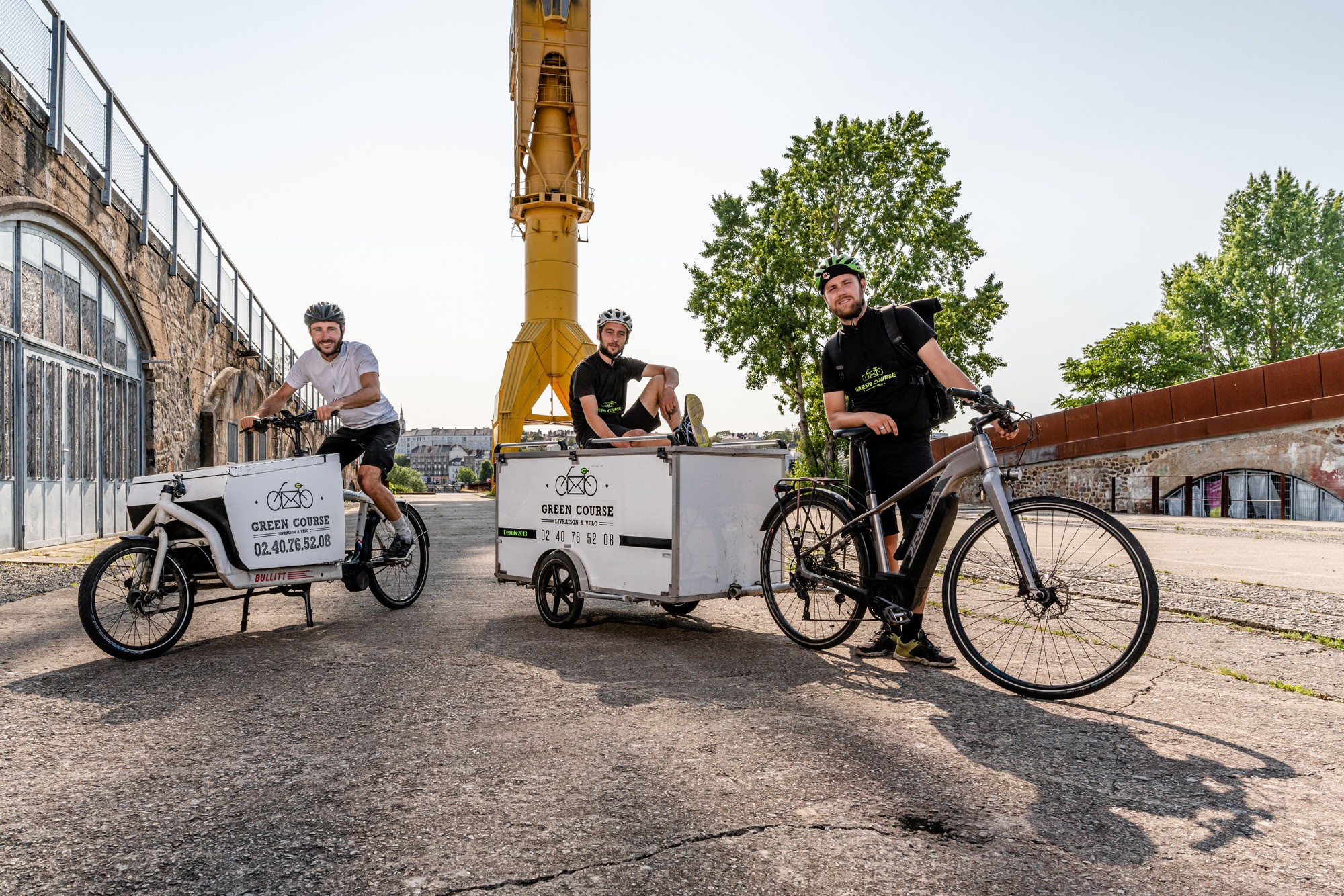 coursier avec un vélo biporteur à plateau chargé de colis. Il est pied à terre, sourit et tient un smartphone à la main.